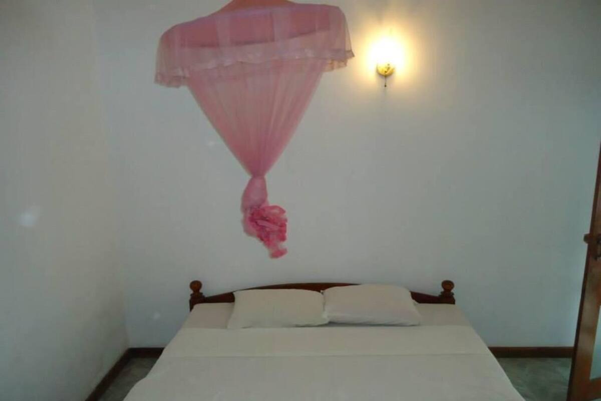 A bed with a wooden headboard and white bedding, a wall-mounted light fixture, and a hanging pink canopy.