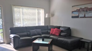 Ein Wohnzimmer mit einem schwarzen Ledersofa, einem Glastisch, einem roten Kissen, einer Stehlampe und einem Gemälde an der Wand.