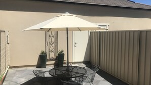 Eine Terrasse mit einem runden Metalltisch und Stühlen, einem weißen Sonnenschirm und zwei Blumentöpfen.