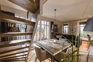 A modern interior with a wooden staircase, a dining area with a table and chairs, and a sofa in a bright room with large windows.