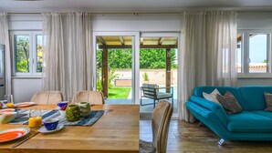 A dining area with a wooden table set for breakfast, a teal sofa, and a view of a garden through glass doors. - Luxurious home in Medulin (Medulin)