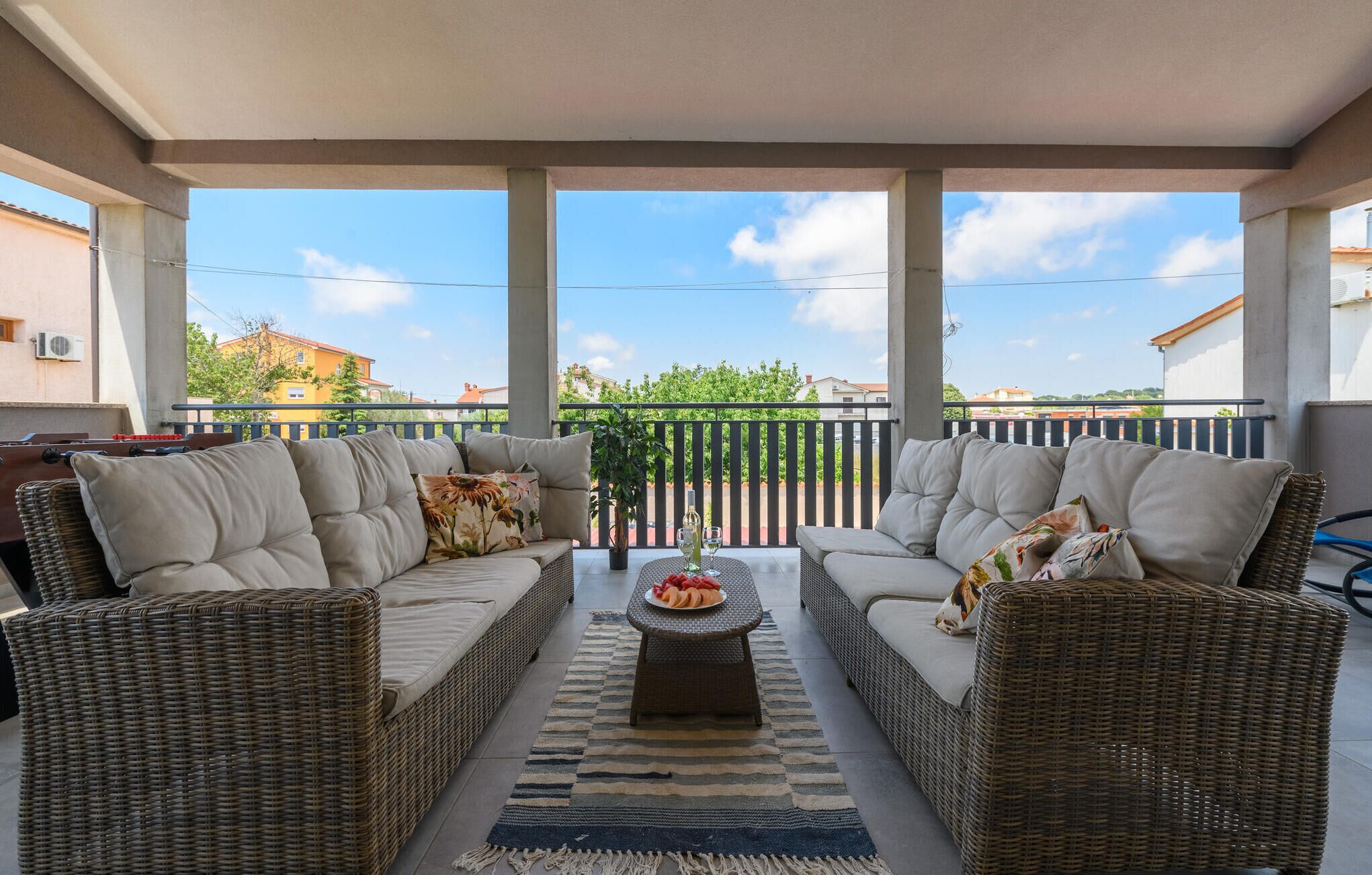 A covered outdoor seating area with wicker furniture and cushions, a central table with a bottle and glasses, and a view of the neighborhood.
