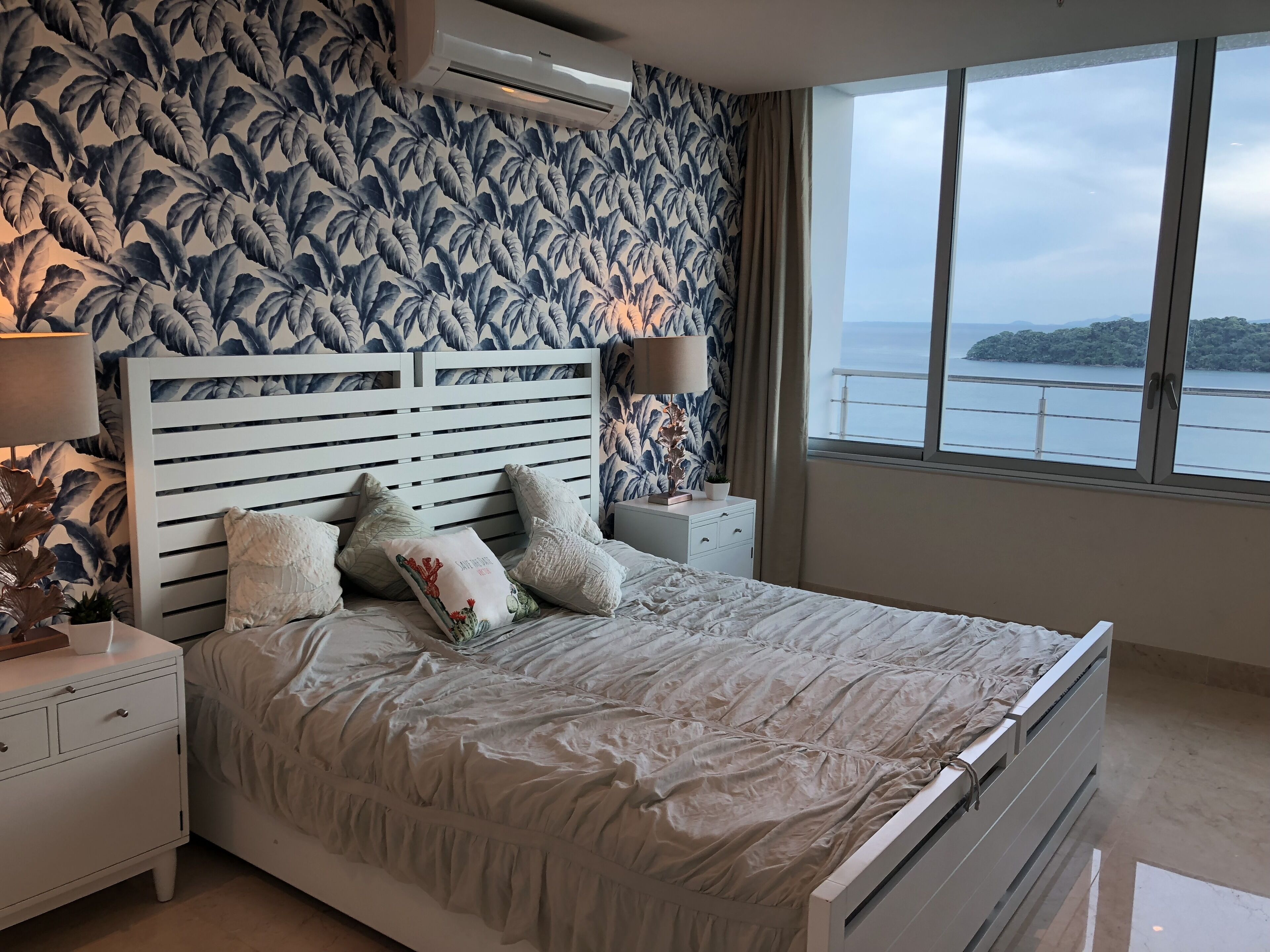 Une chambre à coucher avec un lit, des tables de chevet, une lampe, une plante en pot et une vue sur la mer.