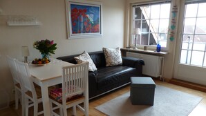 A modern living room with a black leather sofa, white chairs, a dining table with fruit, a grey ottoman, a painting, and a window with blinds. - *'CLOSE TO THE BEACH*'ALLERGY FRIENDLY**MAISONETTE APARTMENT**UP TO 4 PERSONS**BALCONY*** (Friedrichskoog-Spitze)