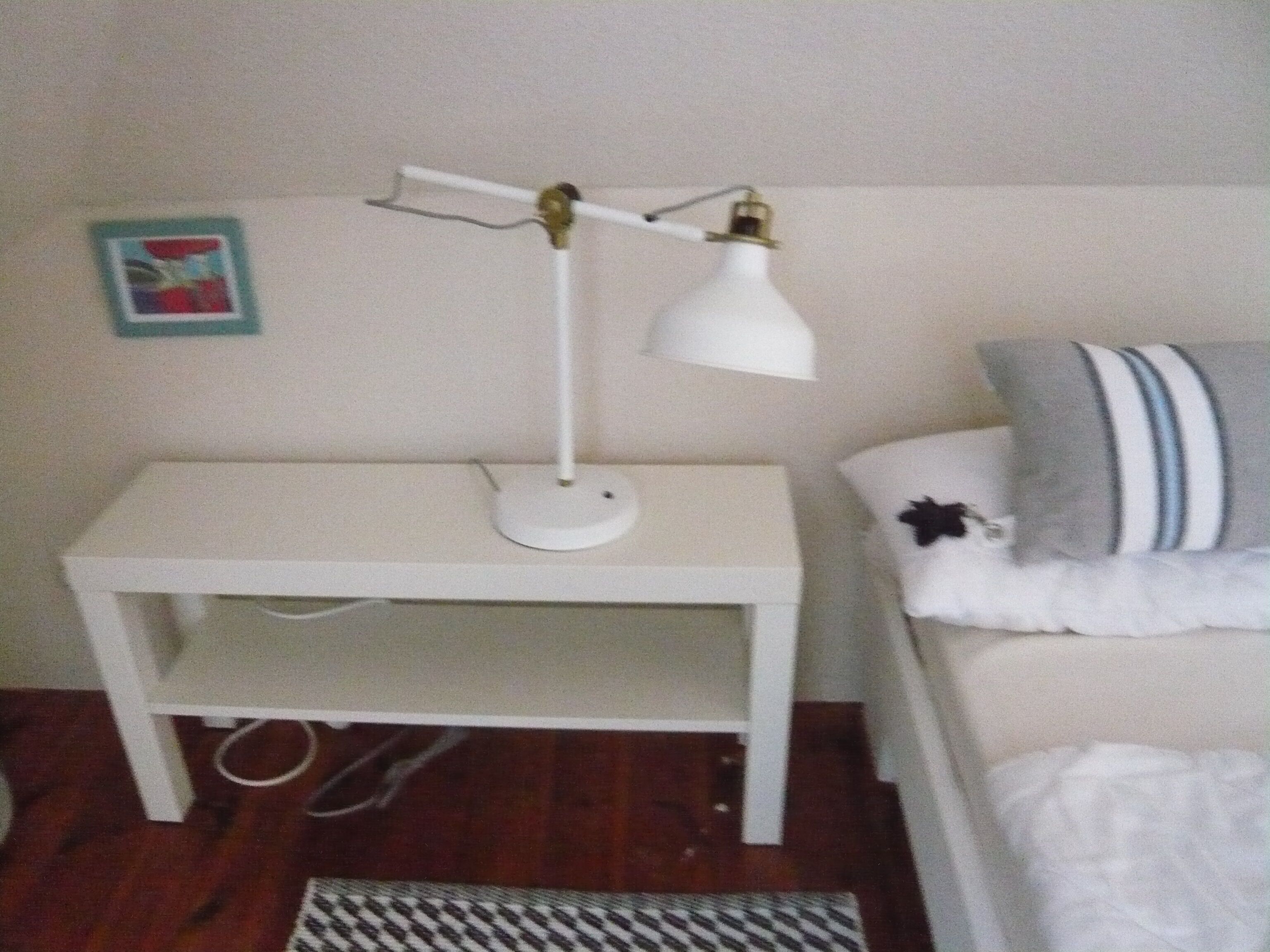 Een klein wit nachtkastje met een witte bureaulamp, een tapijt met dessin en een bed met een gestreept kussen.