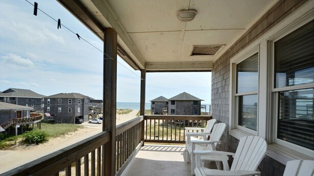 A covered balcony with white chairs, overlooking a beach and buildings. - Pelican Place Cottage- Short walk to beach- Community Pool & Spa (Buxton)
