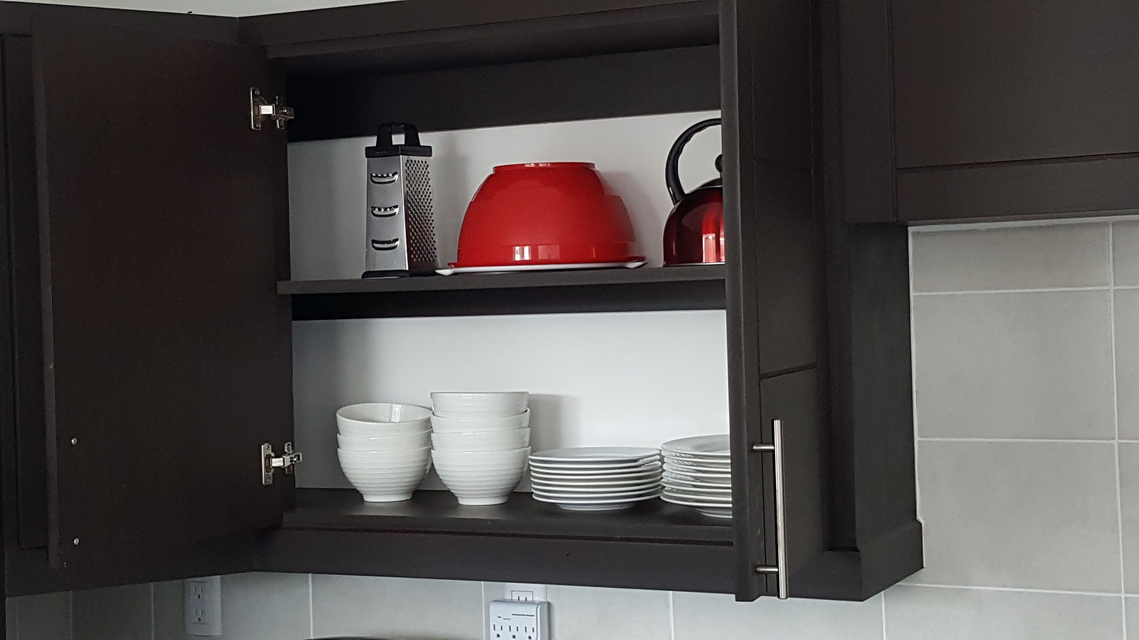 A kitchen cabinet with shelves holding white bowls, plates, a red kettle, and a grater.
