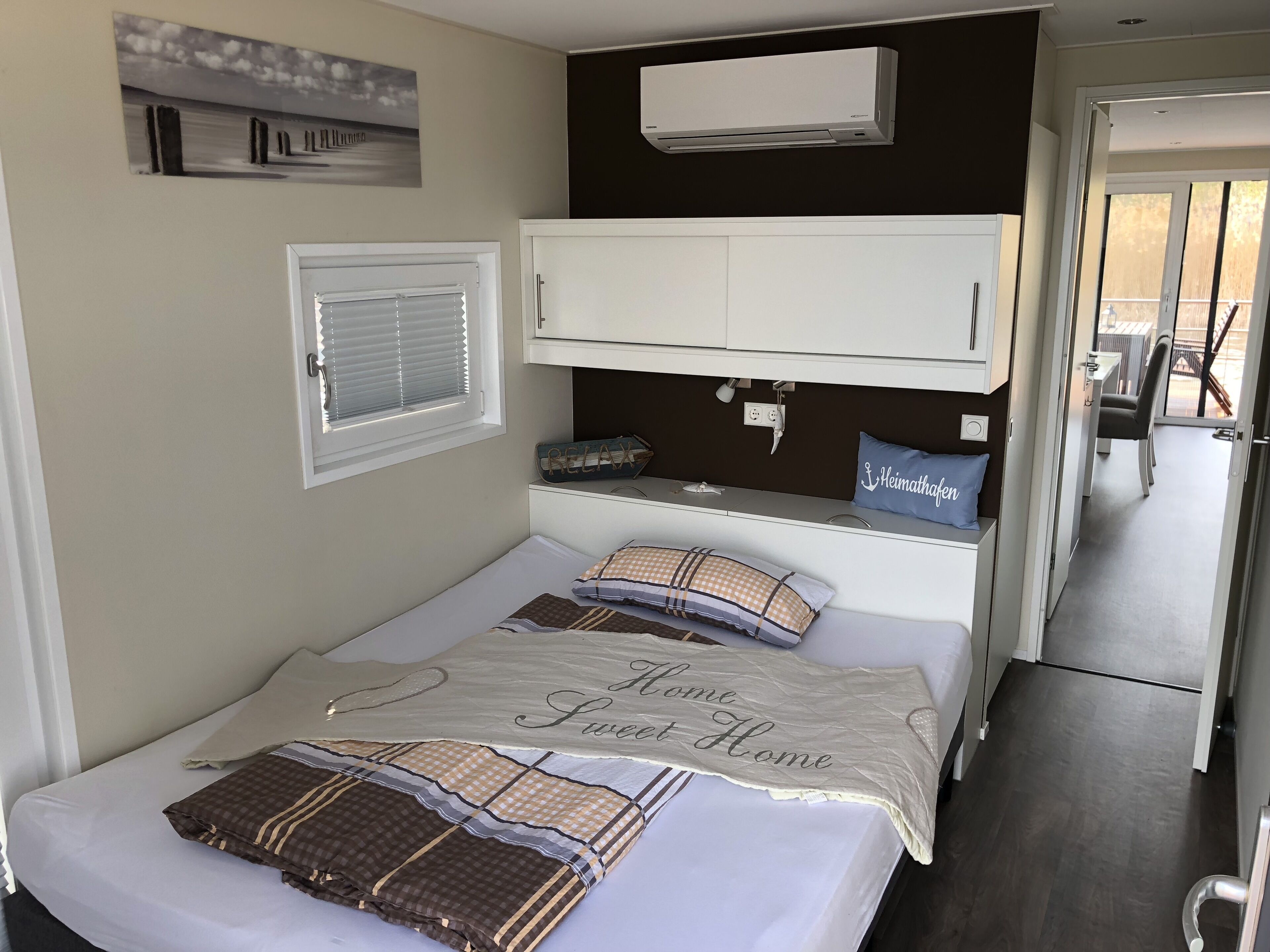 A bedroom with a bed, a white wardrobe, a window with blinds, a wall-mounted air conditioner, and a view of the outside.