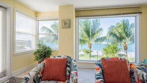 Een kamer met twee fauteuils met bloemenprint, een groot raam met uitzicht op palmbomen en de zee, en een plafondventilator.