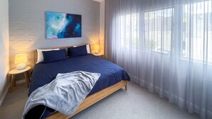 Un dormitorio con una cama grande, una mesita de noche con lámpara, un cuadro en la pared y una ventana con cortinas.
