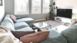 Un salón con un sofá modular, una mesa de centro, un televisor de pantalla plana y ventanas grandes con persianas.