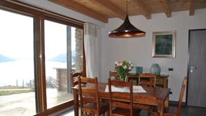 A dining area with a wooden table and chairs, a vase with flowers, a pendant light, a framed picture on the wall, and a view of a body of water through large windows.
