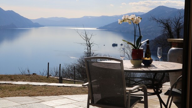A table with a vase of flowers, a wine glass, and a bottle, overlooking a calm lake with mountains in the background.