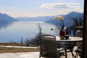 Una mesa con un florero, una copa de vino y una botella, con vistas a un lago tranquilo y montañas al fondo.