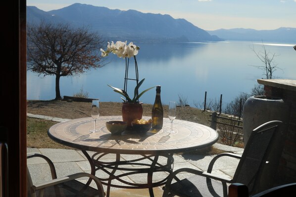Una mesa redonda con una orquídea en maceta, un bol de patatas fritas y una botella de vino, con vistas a un lago tranquilo y montañas al fondo.