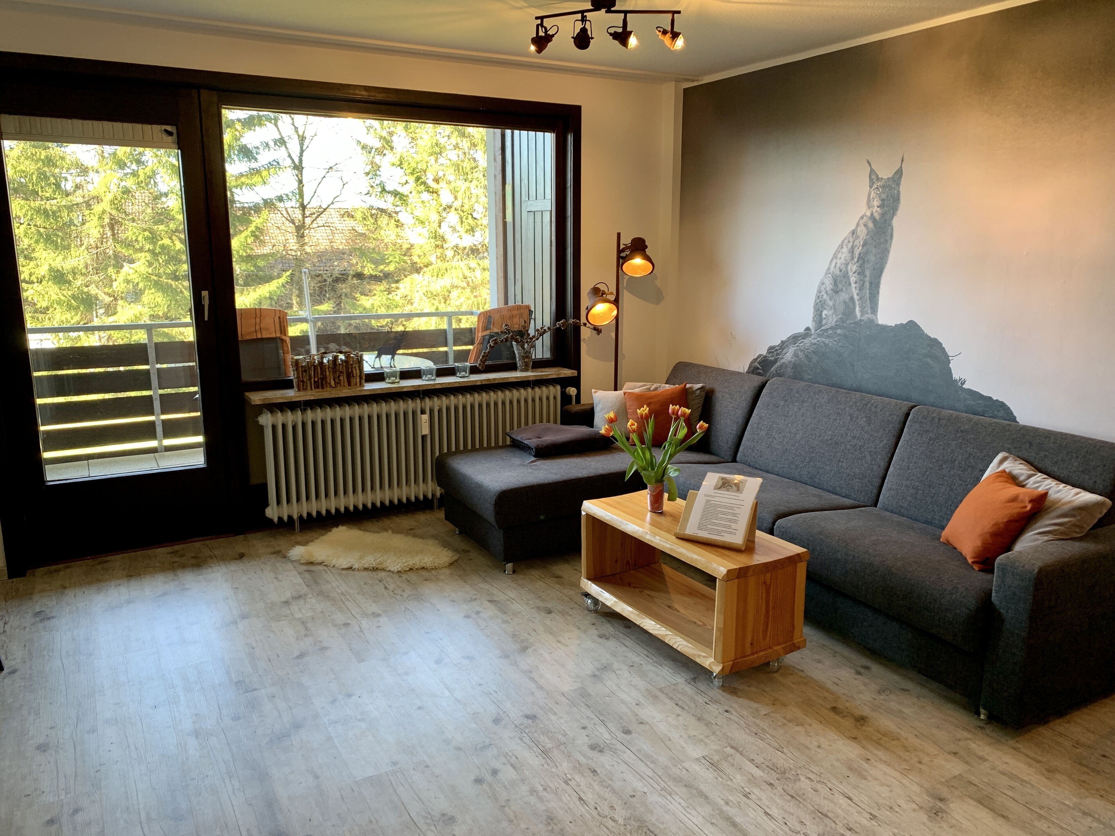 A modern living room with a grey sofa, a wooden coffee table with a vase of flowers, a large window with a view of trees, and a leopard mural on the wall.