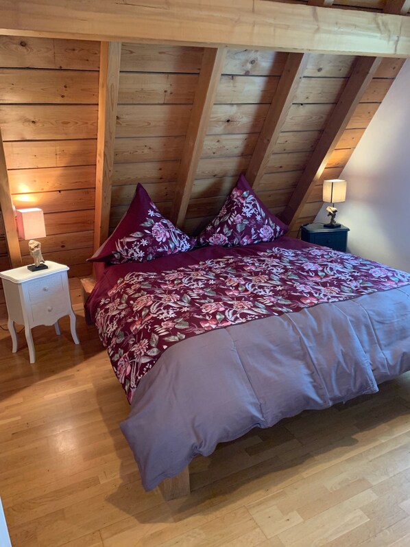 Un dormitorio con techo de madera, una cama con colcha estampada, dos almohadas, una mesita de noche y una lámpara.