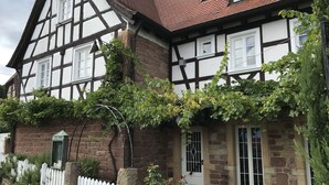 Une maison traditionnelle à colombages, avec une clôture en bois peinte en blanc, des murs en pierre et un toit recouvert de tuiles rouges.