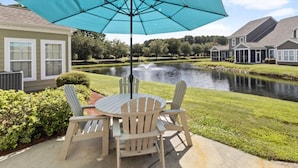 A patio with a table and chairs under a large umbrella, overlooking a pond and houses. - Egret Run #214 Beautiful Condo (North Myrtle Beach)