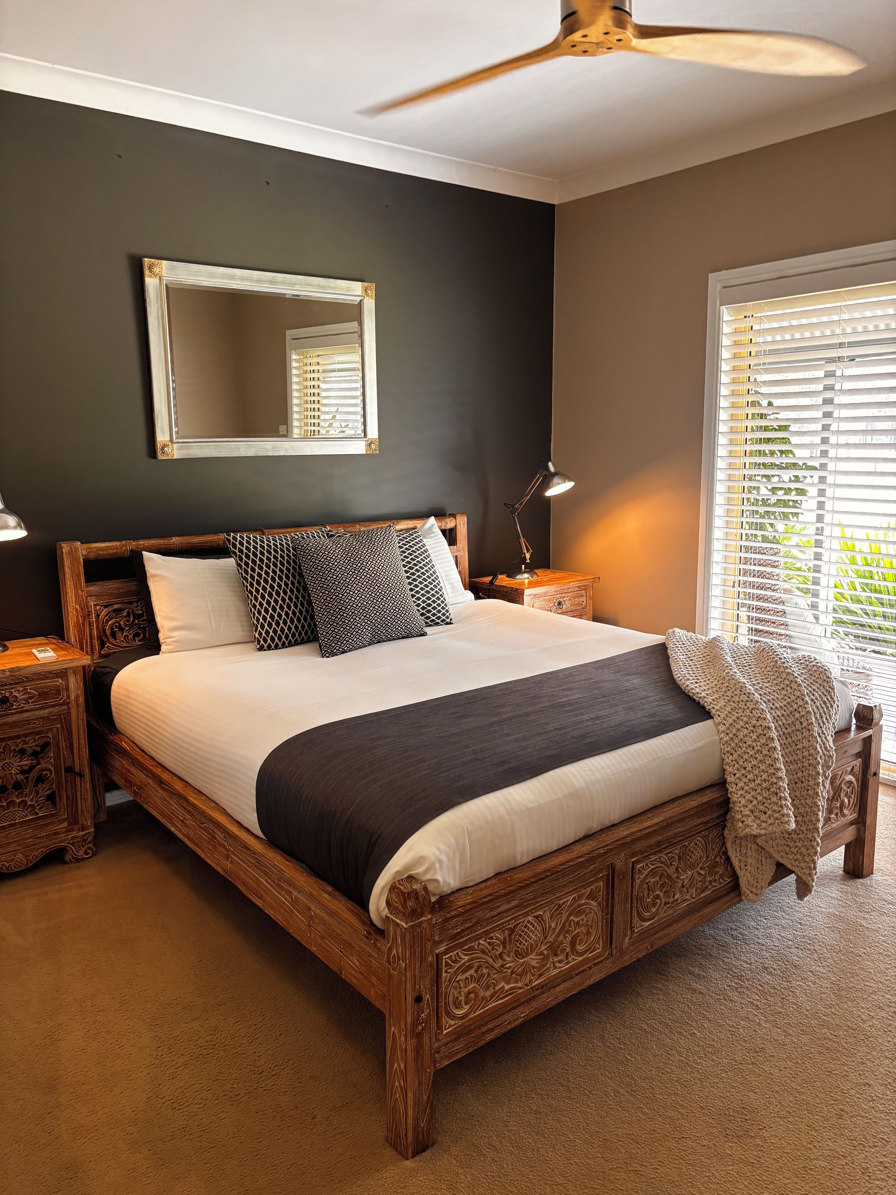 A bedroom with a large bed, a ceiling fan, a bedside table with a lamp, and a window with blinds.