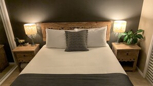 Una cama bien hecha con cabecera de madera, dos mesitas de noche con lámparas y una planta en maceta.