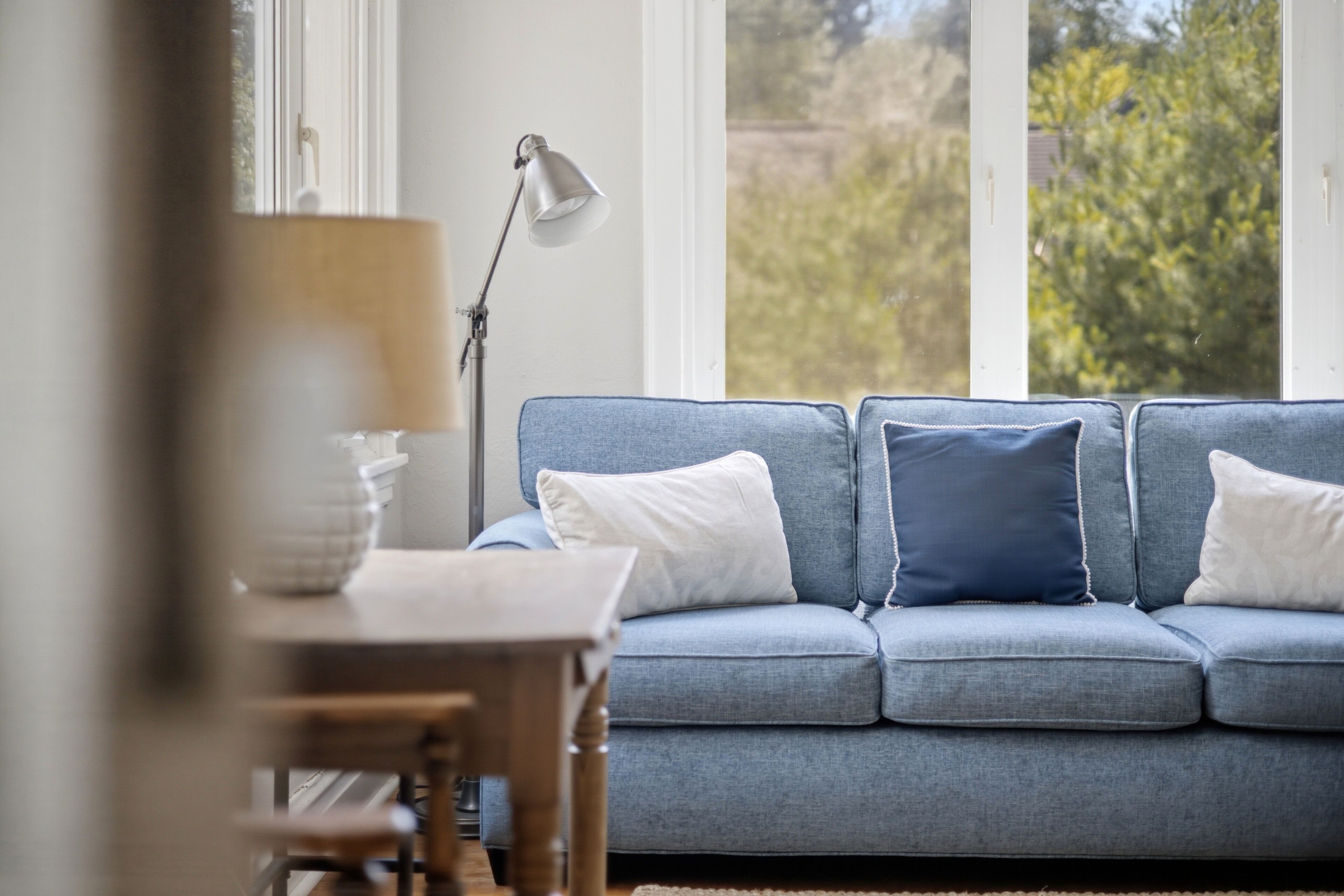 A room with a blue sofa, a lamp, and a view of greenery outside.