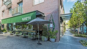Een terras met tafels en stoelen, een groen luifel met de tekst 'MARTA' en bloempotten.