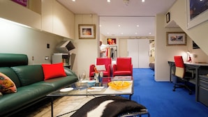 Een moderne woonkamer met een groene bank, rode stoelen, een glazen salontafel en een bureau met stoel. Er hangen ingelijste foto's aan de muur en een trap die naar een andere kamer leidt.