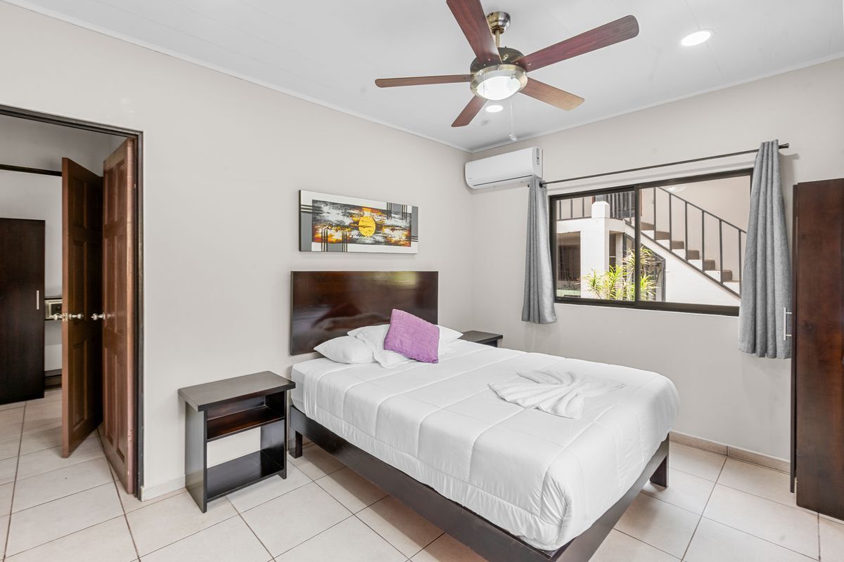 A bedroom with a bed, a ceiling fan, a painting, an air conditioning unit, and a window with a view of a staircase.