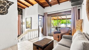 A spacious living area with a sofa, a coffee table, and a chair. A staircase with a balustrade leads to an outdoor area with a view of greenery.