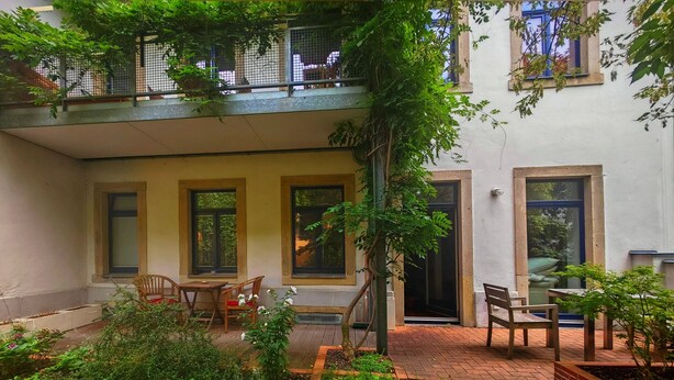 Une cour avec une table et des chaises en bois, un petit jardin et un balcon doté d’une rambarde en métal.