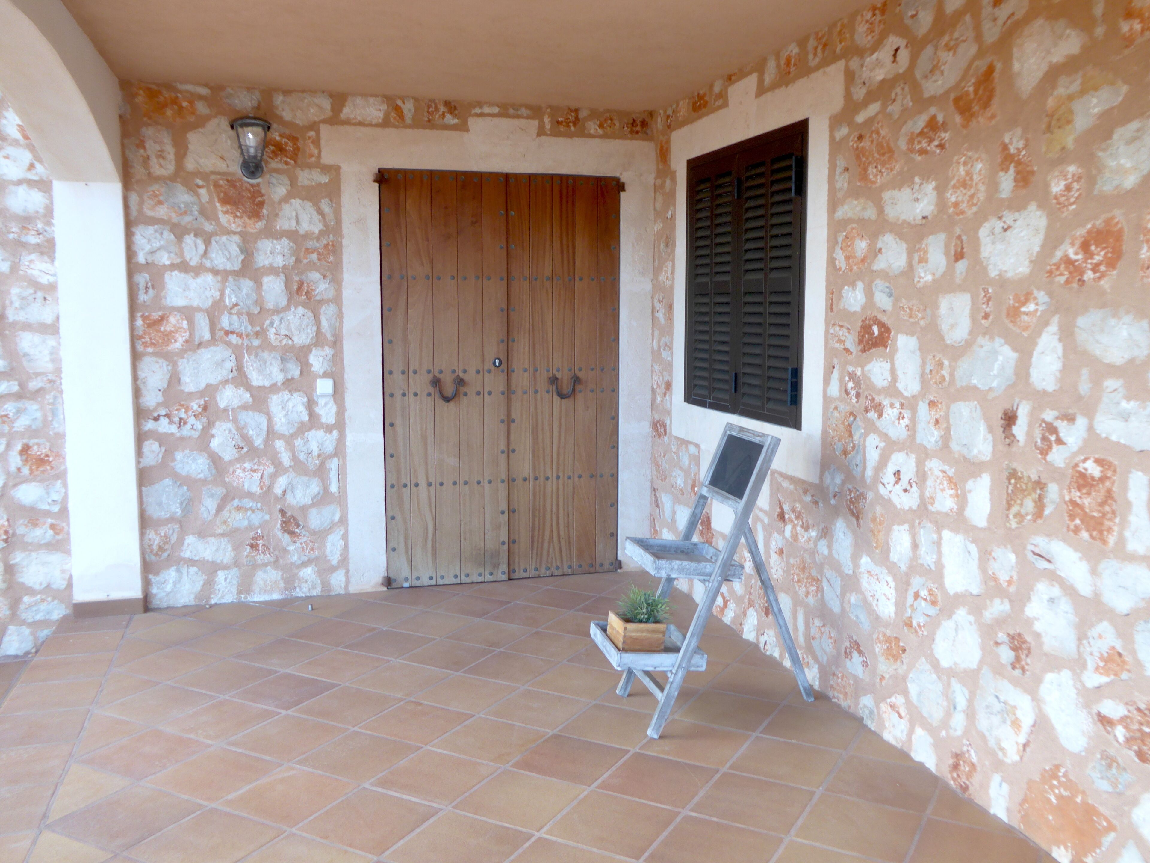 A stone-walled building with a wooden door and window shutters, a tiled floor, and a metal chair with a plant.