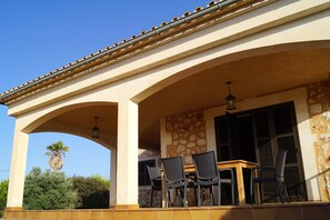Une terrasse couverte avec une table et des chaises, un mur en pierre et un palmier en arrière-plan.