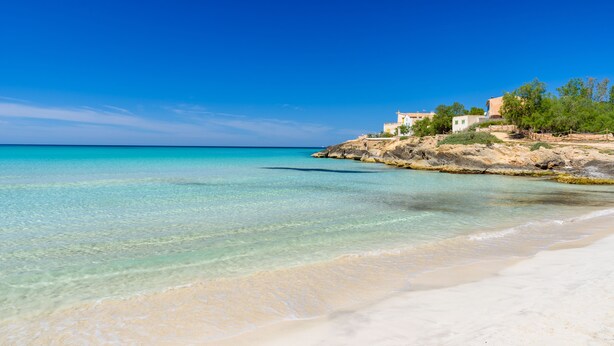 A beach with clear turquoise water, a sandy shore, and a building with a white facade and red roof. - Wonderful finca with a large pool and barbecue kitchen near Es Trenc!  (Campos)