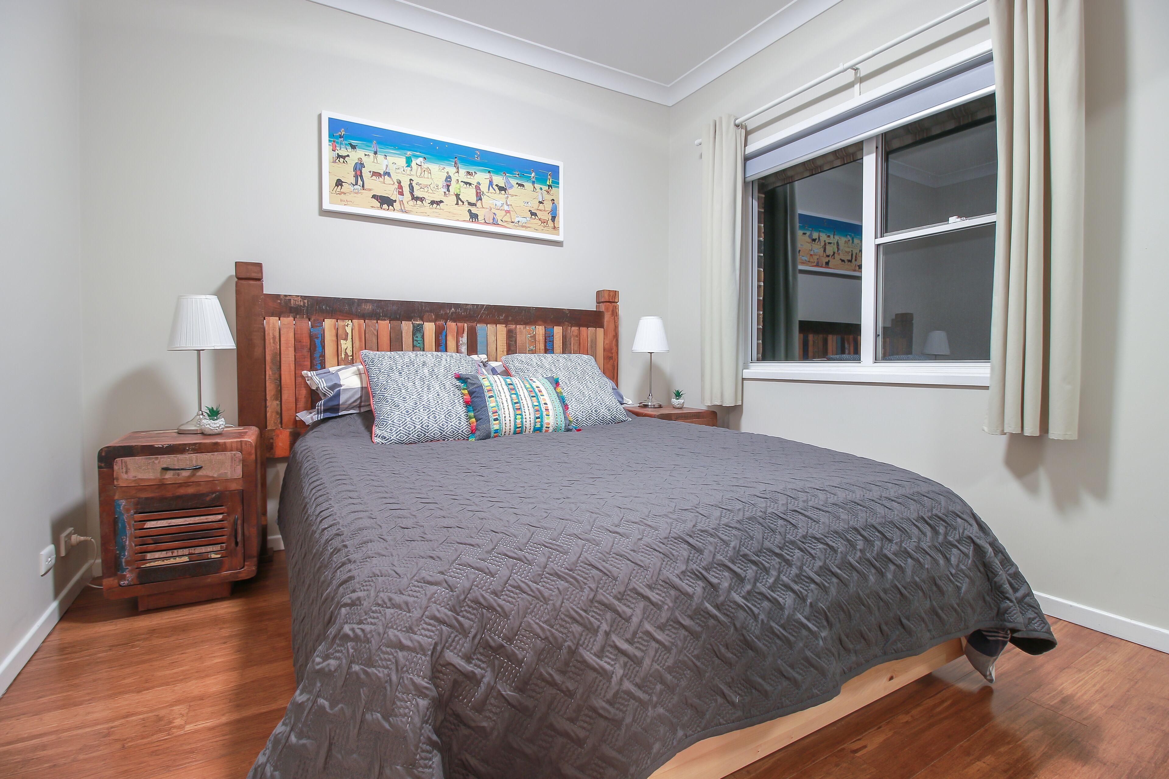A bedroom with a wooden headboard, a grey quilted bedspread, two bedside lamps, a small nightstand, and a window with curtains.
