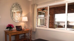A room with a wooden desk, a laptop, a vase with flowers, a mirror, and a window with a view of a brick building and water.