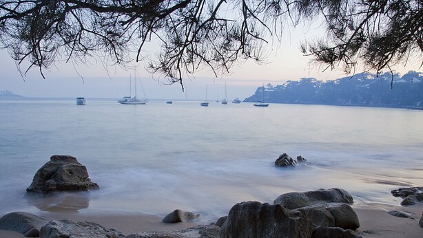 A beach with large rocks in the foreground and boats in the water.
