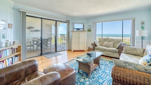 A spacious living room with a large sliding glass door leading to a balcony, a comfortable sofa, a glass coffee table, and a bookshelf filled with books. - CRC 1302 - Beautiful Oceanfront Condo with Amazing Views (St Augustine)
