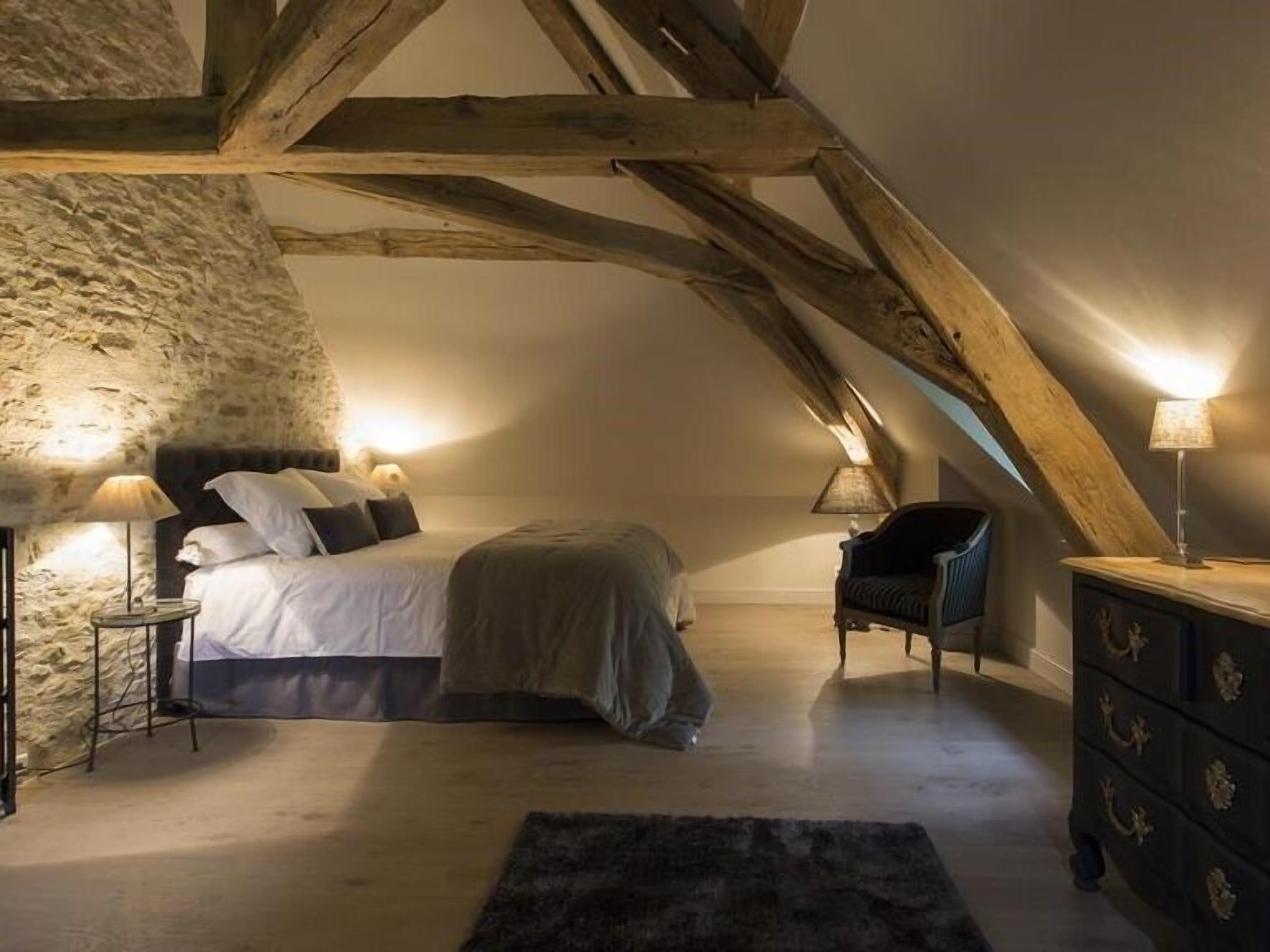 A cozy attic bedroom with exposed wooden beams, a stone wall, a bed with pillows, a chair, and a dresser.