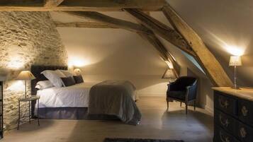 A cozy attic bedroom with exposed wooden beams, a stone wall, a bed with pillows, a chair, and a dresser.