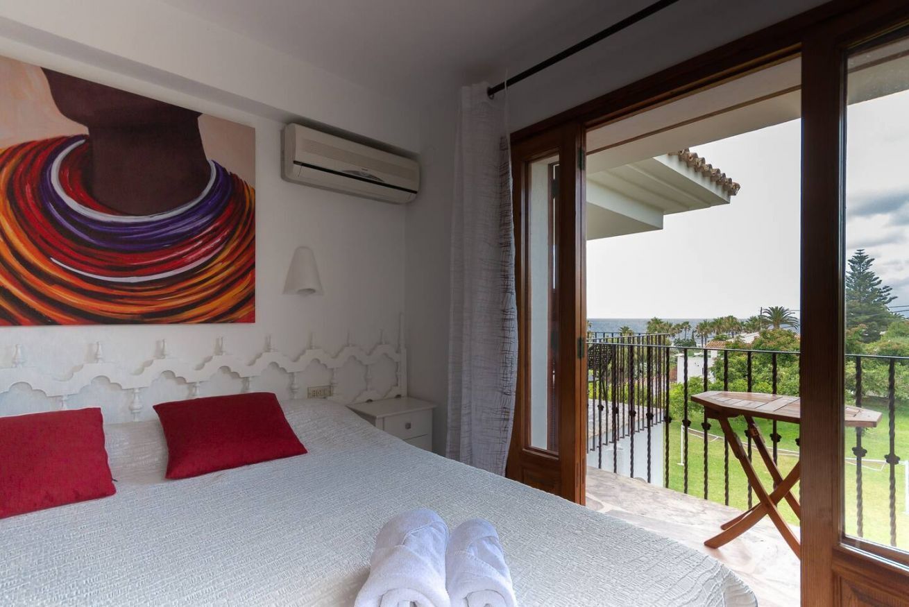 A hotel room with a bed, red pillows, a painting, an air conditioner, and a balcony with a table and chairs.