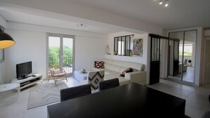 Ein großzügiges Wohnzimmer mit einem großen schwarzen Tisch, einem weißen Sofa und einem Fernseher auf einem weißen Schrank. Ein Balkon mit Stuhl und Tisch ist vorhanden, und eine Glastür verschiebt sich zu einem weiteren Raum.