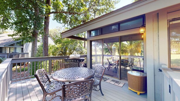 Eine überdachte Terrasse im Freien mit einem runden Tisch und Stühlen, einem Grill und Blick auf die umliegenden Bäume und den Garten.