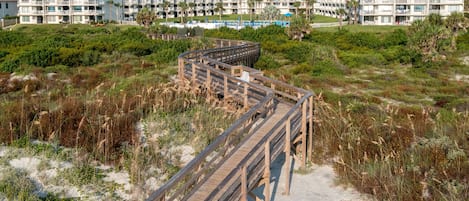 A wooden boardwalk leading to a beachfront hotel with multiple balconies and a pool area.