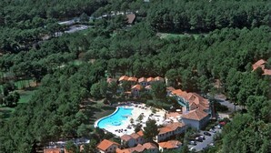 An aerial view of a resort with a central swimming pool, surrounded by villas with red-tiled roofs, set in a wooded area.
