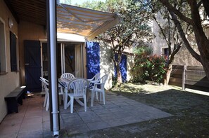 Eine Terrasse mit Tisch und Stühlen, ein überdachter Bereich und ein Garten mit Bäumen und einem Zaun.