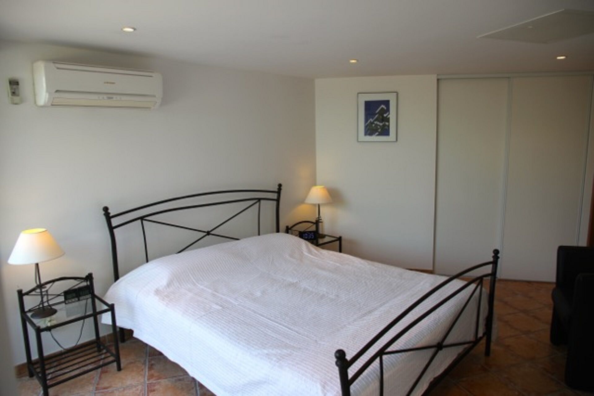 Une chambre à coucher avec un lit, deux tables de chevet avec des lampes, un climatiseur, un tableau encadré au mur et une porte.