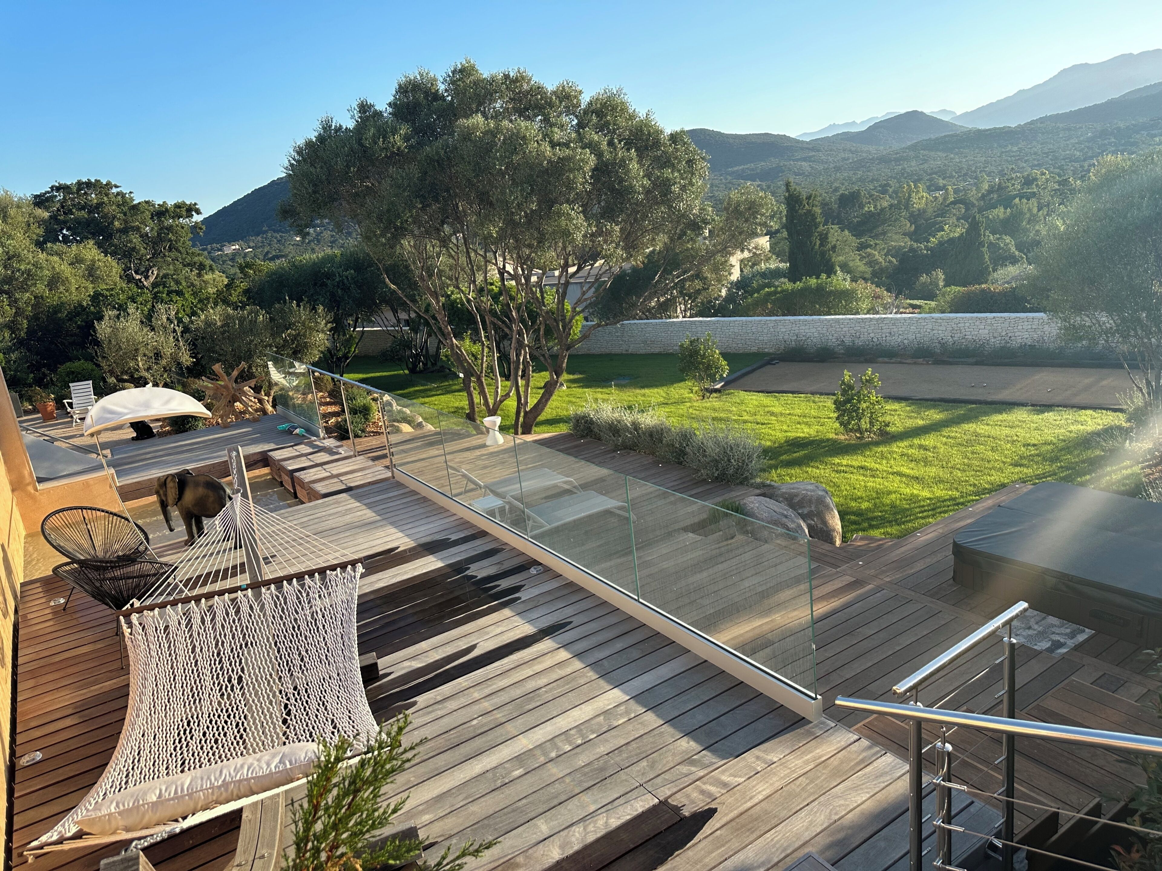 Una terraza de madera con una hamaca, baranda de vidrio y vista a un paisaje verde con árboles y un muro de piedra.
