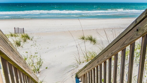 Eine Holztreppe führt zu einem Sandstrand mit dem Meer im Hintergrund.