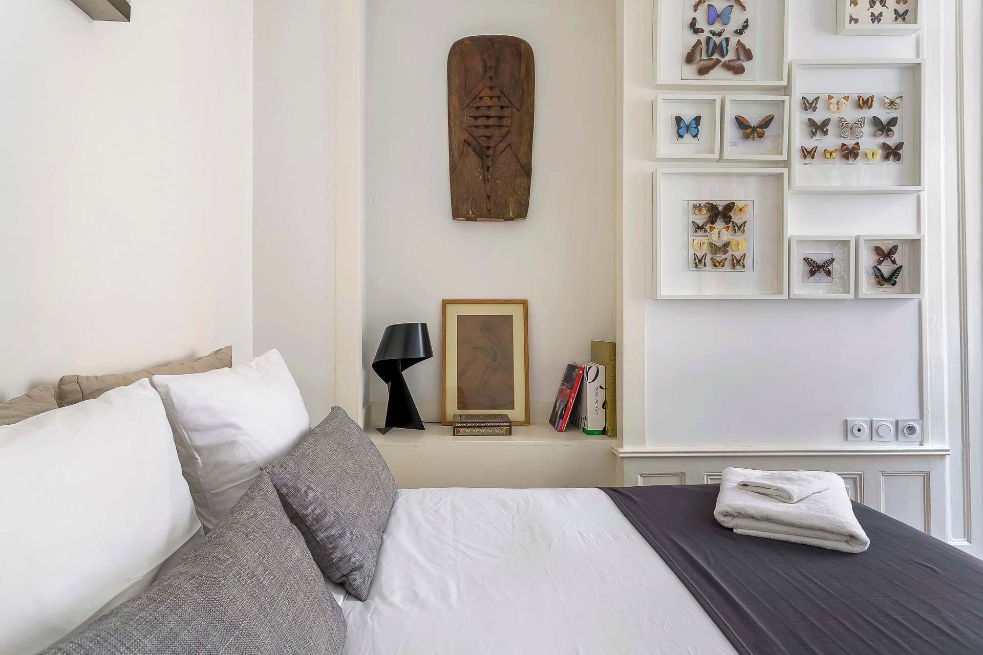 Una cama bien tendida con ropa de cama blanca y gris, una lámpara negra, una obra de arte enmarcada y un estante montado en la pared con adornos de mariposas.